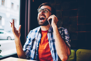 Casual dressed angry bearded young man crying and arguing during phone conversation on smartphone...