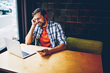 Carefree bearded young man singing song while listening audio in modern headphones connected via...