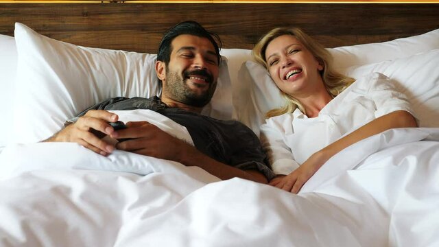 Couple watching television together on cozy bed on weekend holiday. Handsome husband and beautiful wife love to stay together and enjoy doing activity together. They are lovely couple and just marry