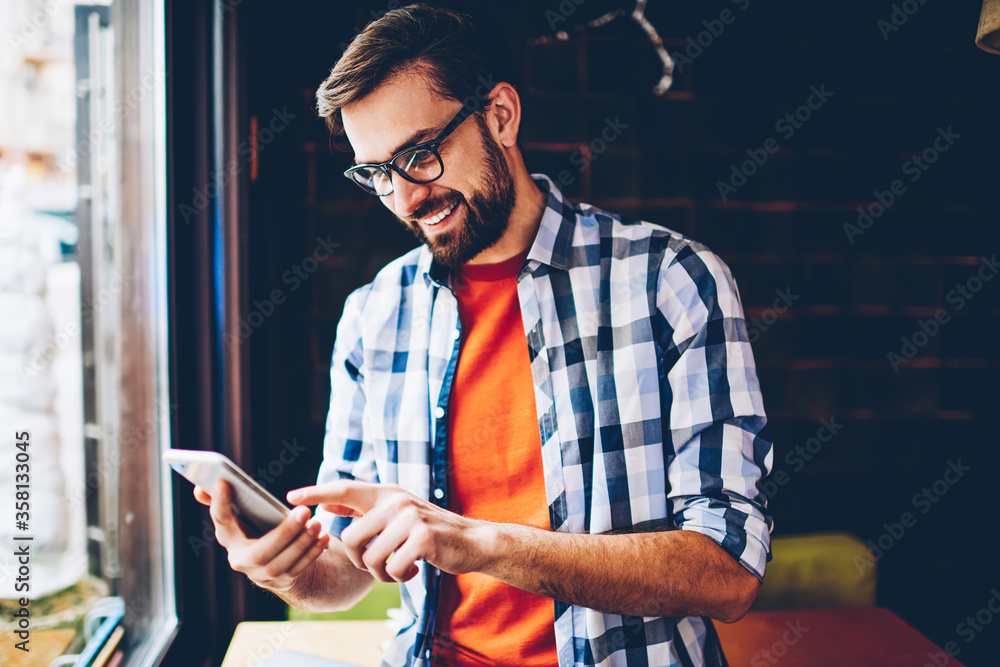 Poster cheerful bearded blogger laughing while reading funny comments in social networks on smartphone devi