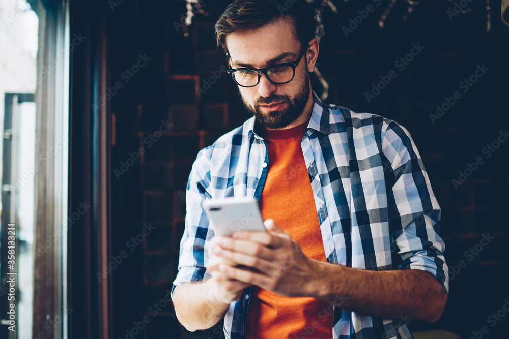 Poster pensive bearded hipster blogger reading incoming notification and chatting in networks on smartphone