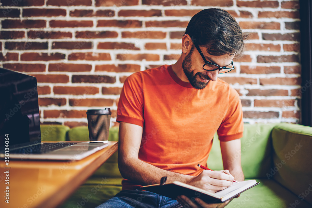 Aufkleber cheerful bearded student writing down notes of course work in notepad sitting in coworking space at 
