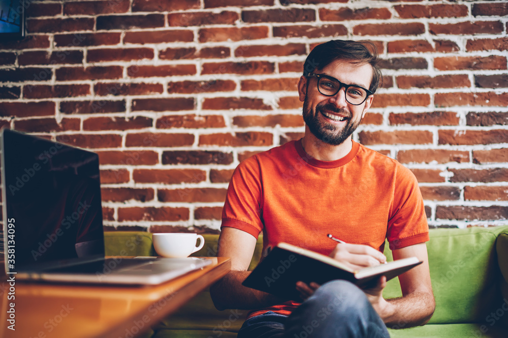 Aufkleber half length portrait of successful bearded young man in eyewear smiling at camera while writing down