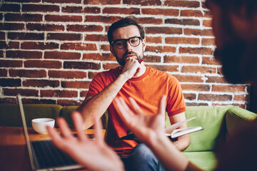 Attentive bearded male employer listening creative ideas from professional during meeting sitting at laptop computer in stylish coworking space.Pondering young man discussing ideas for own project