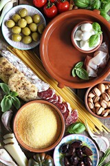 Italian dinner table concept with salami, vegetables variety, pasta, polenta, cheese, nuts and herbs. Ingredients for cooking of traditional mediterranean dinner or lunch. Overhead view