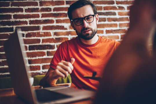 Sceptical bearded young man in optical eyeglasses looking at modern laptop computer device connected to 4G internet and thinking on technical problem sitting in coworking space interior