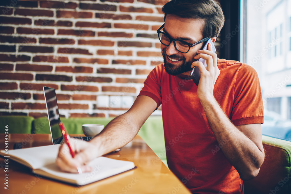 Poster cheerful bearded young man calling on smartphone device and writing text information in notepad sitt