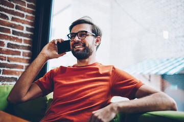 Positive hipster guy with beard calling on smartphone device to arrange meeting with friend.Joyful young man in stylish eyeglasses talking on modern mobile phone resting in coffee shop interior