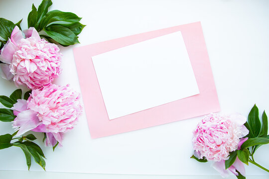 Creative layout of pink peonies flowers on a white background with a paper card for text. Flat lay, minimalism and nature concept.