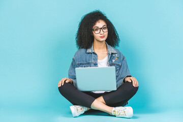 Portrait of smiling young afro american woman using laptop while sitting on a floor with legs crossed isolated over blue background.