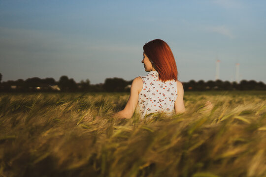 Red-haired girl stands in a field of grain turned back to photograph. In the background the blue sky