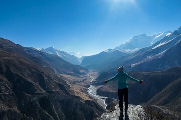 Obraz premium A woman wearing a beanie and blue jumper, spreads her arms wide, breathing deeply the fresh mountain air. Freedom and happiness. Below Manang valley stretches in Himalayas, along Annapurna Circuit.