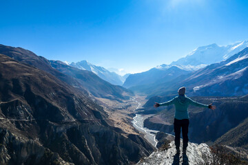 A woman wearing a beanie and blue jumper, spreads her arms wide, breathing deeply the fresh mountain air. Freedom and happiness. Below Manang valley stretches in Himalayas, along Annapurna Circuit.