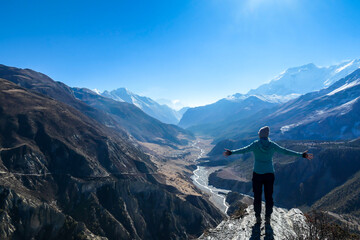 A woman wearing a beanie and blue jumper, spreads her arms wide, breathing deeply the fresh...