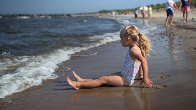Sea waves water washing legs of lovely little girl sitting on coastline shore