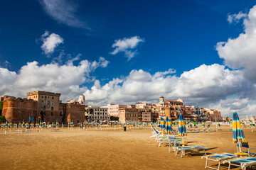 The closed umbrellas, the empty sunbeds and deckchairs and the deserted beach in a bathhouse, on a windy day in late summer. The cloudy blue sky. Nobody, no people. Nettuno, Rome, Lazio, Italy.