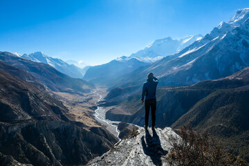 A man wearing a beanie and blue jumper, stands at the rock edge, breathing deeply the fresh mountain air. Freedom and happiness. Below Manang valley stretches in Himalayas, along Annapurna Circuit.