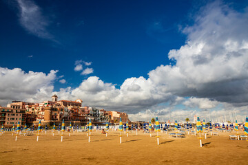 The closed umbrellas, the empty sunbeds and deckchairs and the deserted beach in a bathhouse, on a windy day in late summer. The cloudy blue sky. Nobody, no people. Nettuno, Rome, Lazio, Italy.