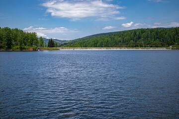 Fototapeta premium Beautiful mountain lake with wavy blue water with reflection and green forest and white fluffy cloud sky on summer day. Travel, tourism, and resort in nature concept. Vlasina lake, Serbia, Europe.