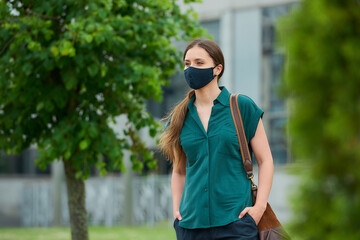 Naklejka premium Woman in medical face mask walks between trees thrusting hand into pocket of trousers, holds a bag in downtown. Girl keeping social distance wears a protective mask to avoid the spread of coronavirus