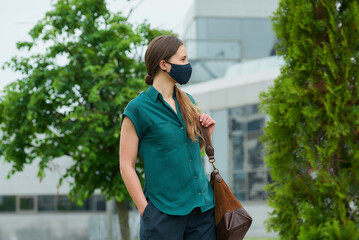 A woman in a medical face mask walks between trees thrusting a hand into a pocket of trousers, holds a leather bag and looks to the side in downtown. A girl in protective mask keeping social distance