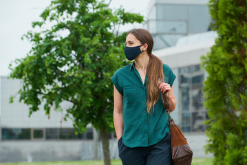 Woman in medical face mask walks near trees thrusting hand into pocket of trousers, holds bag in downtown. Girl keeping social distance wears a protective face mask to avoid the spread of coronavirus