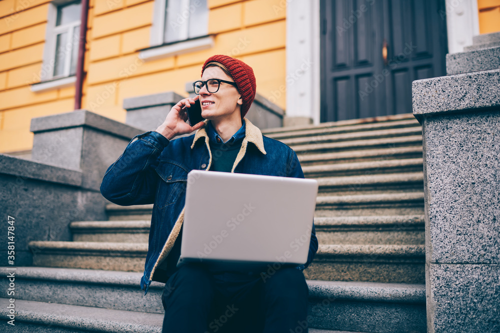 Poster positive hipster blogger calling on smartphone device and laughing during friendly conversation whil