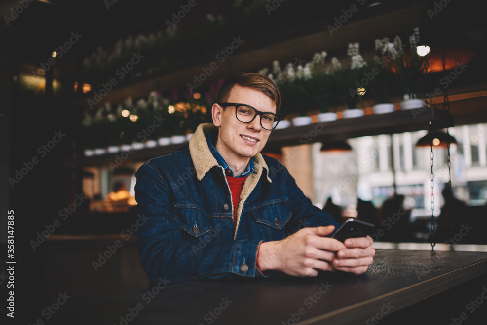 Poster half length portrait of successful male blogger in denim jacket smiling at camera while updating pro