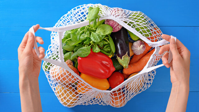 hands holding string bag with vegetables on blue background, eco shopping, top view