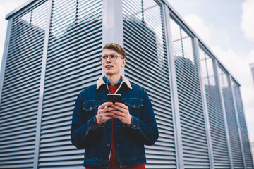 Fototapeta premium Pondering young male blogger in stylish denim jacket looking away while messaging in chat on smartphone using 4G internet standing in downtown.Contemplative hipster guy installing app on cellular