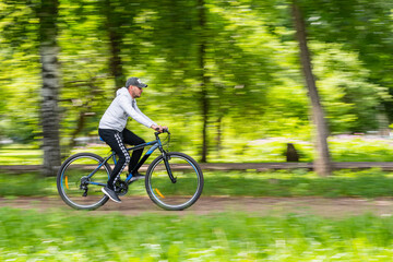 A young guy rides a bicycle at high speed on a treadmill of a school stadium against the background of an apple orchard. Blurred abstract green background.