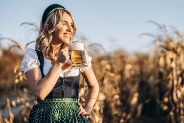 Pretty happy blonde in dirndl, traditional festival dress, holding mug of beer outdoors in the field