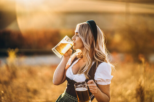 Pretty happy blonde in dirndl, traditional festival dress, holding two mugs of beer outdoors in the field