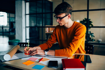 Pensive male engineer drafting sitting at working place with equipment making notes of ideas on...