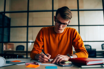 Thoughtful male designer concentrated in making plan for productive process on project sitting in office, skilled architect drawing sketch and checking accountings using stationery and equipment