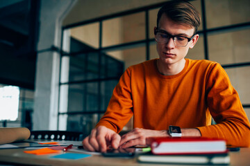 Young student making researches and accountings creating coursework sitting at desktop with stationery, professional serious male architect concentrated on project during  working process in office