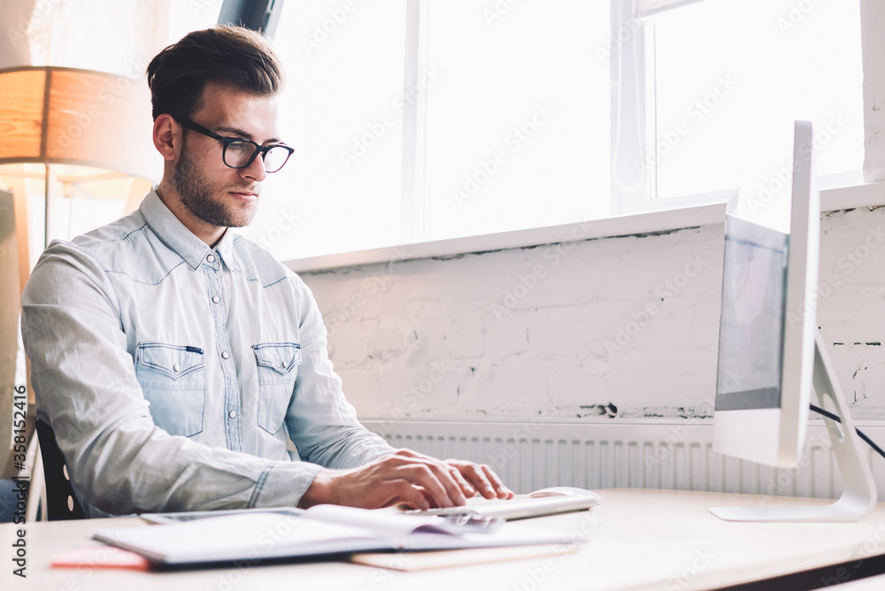 Poster serious male software developer keyboarding program code on modern computer sitting at office deskto