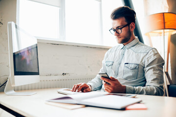 Concentrated young IT developer checking mail on smartphone while working in office on computer,...