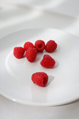 The sun shines on the fresh ripe raspberries on the white plate. Light and shadow on white background.