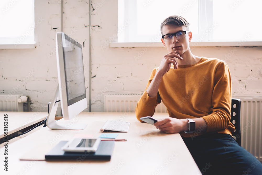 Poster confident male software developer in spectacles looking at camera holding smartphone device and wait