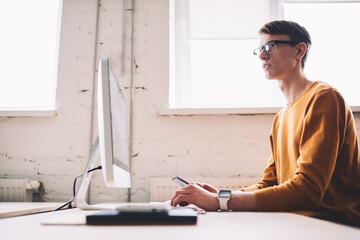 Thoughtful caucasian male it developer pondering on creating website sitting with modern computer on front, young contemplative man in spectacles for vision correction using technology for working