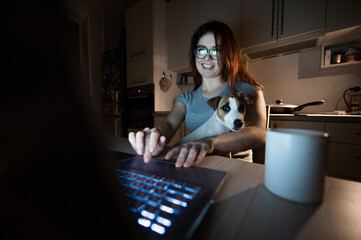 A smiling woman in glasses sits at a wireless computer in the kitchen with a puppy of Jack Russell Terrier on her knees. Girl freelancer works at a laptop at home and drinks coffee.