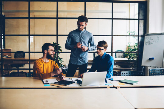 Crew of male IT developers presenting to young CEO programs on modern laptop during meeting, professional team of designers having brainstorming session discussing ideas and solutions together
