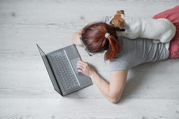 A European red-haired woman is lying on the floor with her dog and is typing on a laptop. Female...
