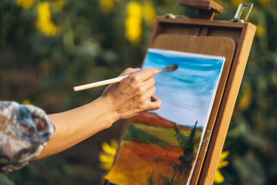 Close up hands of female artist holding brush and draws a picture of the field. Blurred background with easel in sunflower field