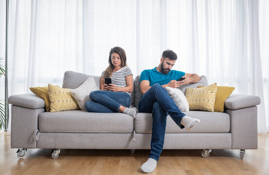 Young Couple Sitting On The Sofa In The Living Room And Ignoring Each Other By Surfing And Text Messaging On Their Smartphones, Relationship Difficulties And Issues Concept