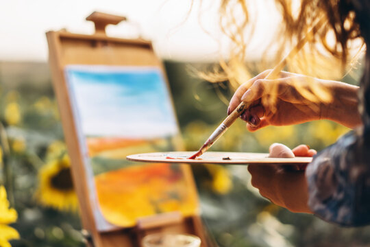 Close up hands of female artist holding brush and palette with oil paints. Blurred background with easel in sunflower field