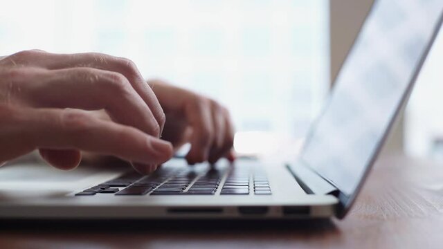 Close-up of hands of unrecognizable businessman typing on laptop computer at the desk in modern room on background of large window. Concept of remote home working. Shooting in slow motion.