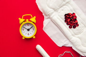 Obraz na płótnie Canvas Close-up of feminine pad and cotton tampon with red glitters and yellow alarm clock on red background.