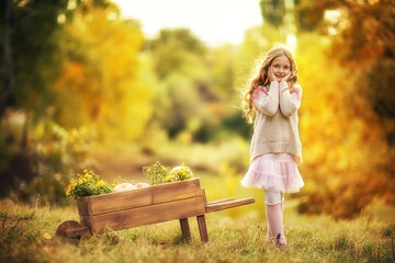 Young girl in sweater and pink gaiters posing near a wooden wheel-barrow with pumpkins. Fall...